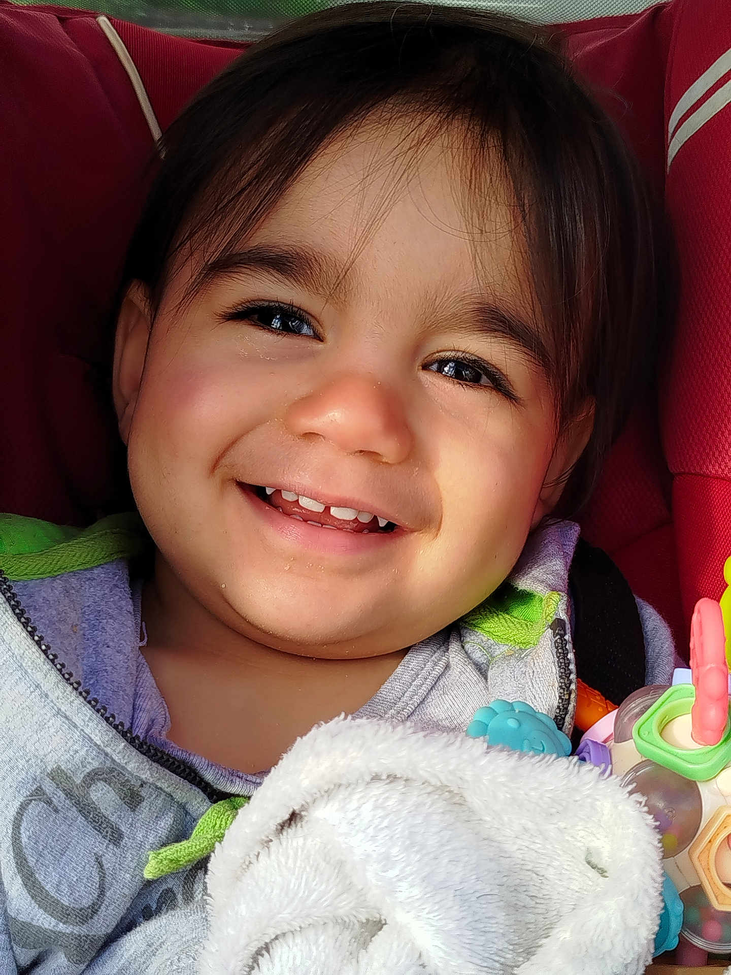 Orion participe au concours pour gagner de l'argent avec cette photo : child, toddler, smile, teeth, face, eyes, dark_hair, cheeks, car_seat, red_seat, blanket, plush, toy, rattle, hoodie, closeup, portrait, happy, candid, sunlight