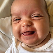 Bayleigh is registered to the contest to win money with this photo: baby, blanket, cheeks, child, close_up, cute, expression, face, happy, indoors, infant, lying_down, mouth, person, portrait, skin, smiling, tongue, white_shirt, young