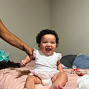 Soleia joined the competition — help win amazing prizes! baby, child, smiling, curly_hair, bed, white_clothing, hand, adult, support, indoor, room, pillow, blanket, person, infant, happy, seated, skin, foot, cute