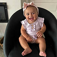 Naiomi is registered to the contest to win money with this photo: baby, child, smiling, headband, bow, pink_clothing, ruffles, chair, black_chair, indoors, happy, cute, sitting, toes, furniture, infant, person, portrait, laughing, young_child