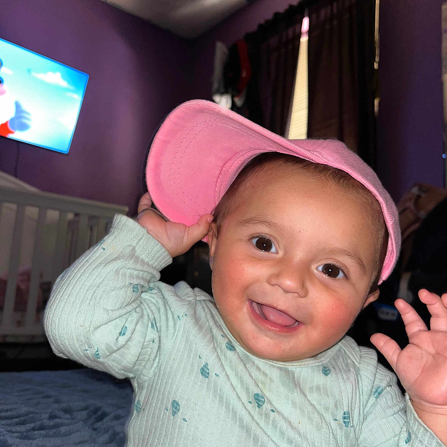 Naiomi joined the competition — help win amazing prizes! baby, bedroom, cartoon, child, crib, curtains, cute, expression, face, hand, happy, indoor, light_green_sweater, person, pink_cap, playful, purple_wall, smiling, television, toddler