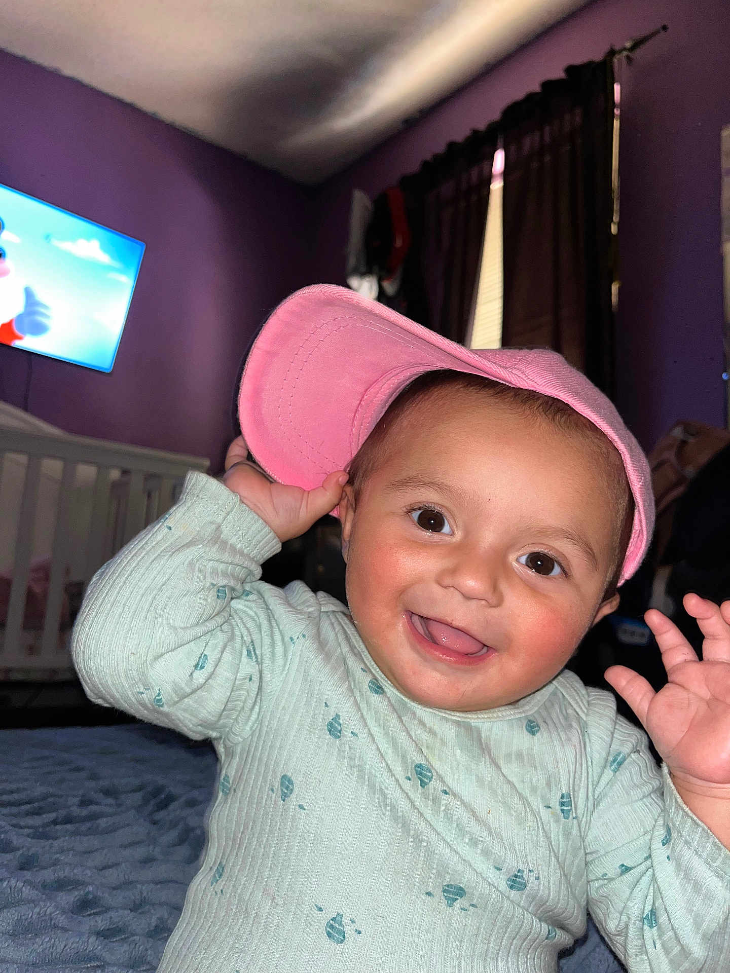 Naiomi joined the competition — help win amazing prizes! toddler, child, smiling, pink_cap, light_green_sweater, indoor, bedroom, crib, television, cartoon, purple_wall, curtains, happy, baby, person, face, hand, expression, cute, playful