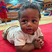 Kadir is registered to the contest to win money with this photo: baby, child, indoor, red_carpet, curly_hair, white_shirt, play_area, background_children, blurry_background, casual_clothing, hands_clasped, floor, candid, cute, expression, portrait, diverse_children, activity, fun, playful