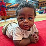 baby, child, indoor, red_carpet, curly_hair, white_shirt, play_area, background_children, blurry_background, casual_clothing, hands_clasped, floor, candid, cute, expression, portrait, diverse_children, activity, fun, playful