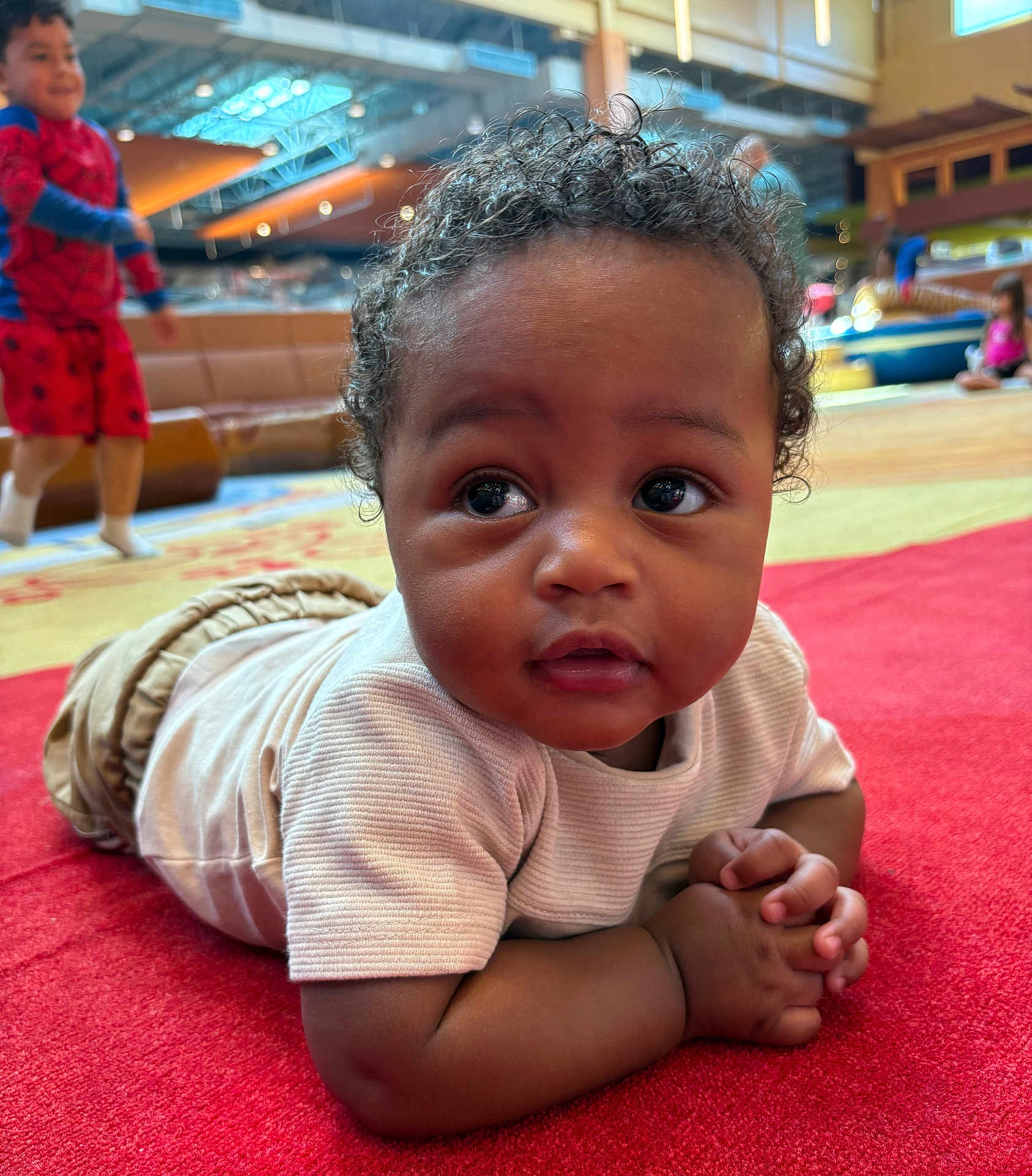 Kadir is registered to the contest to win money with this photo: baby, child, indoor, red_carpet, curly_hair, white_shirt, play_area, background_children, blurry_background, casual_clothing, hands_clasped, floor, candid, cute, expression, portrait, diverse_children, activity, fun, playful