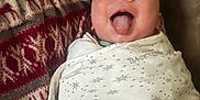 Benson joined the competition — help win amazing prizes! baby, infant, face, tongue, swaddle, blanket, pattern, stars, moon, reindeer, red, beige, cozy, happy, playful, cute, indoor, person, portrait, smile