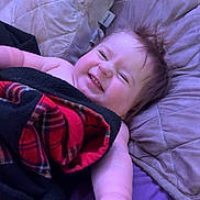 Ivy is registered to the contest to win money with this photo: baby, smiling, blanket, plaid, bed, pillow, happy, child, toddler, face, skin, hair, hand, lying_down, cozy, indoors, cute, expression, relaxing, comfort