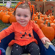 Valerie is registered to the contest to win money with this photo: autumn, black_pants, blue_eyes, child, cute, fall, festive, grass, hair_ribbons, holiday, orange_clothing, outdoor, person, pumpkin, pumpkin_patch, seasonal, smile, toddler, tree, wagon