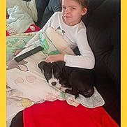 Aïka participe au concours pour gagner de l'argent avec cette photo : child, dog, puppy, blanket, couch, remote_control, indoor, resting, relaxed, smiling, white_shirt, black_dog, curled_up, comfort, home, carpet, pillow, young_girl, pet, casual