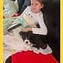 child, dog, puppy, blanket, couch, remote_control, indoor, resting, relaxed, smiling, white_shirt, black_dog, curled_up, comfort, home, carpet, pillow, young_girl, pet, casual
