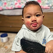 Mataio is registered to the contest to win money with this photo: toddler, child, baby, face, tongue, hairbrush, hand, clothing, indoor, furniture, pillow, rosy_cheeks, expression, person, cute, sitting, floor, blurred_background, smiling, hoodie