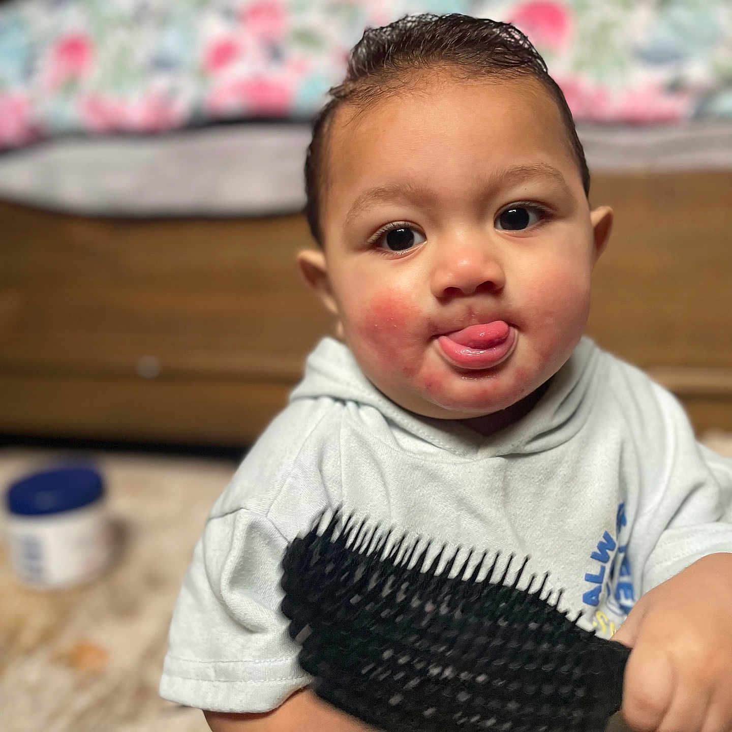 Mataio is registered to the contest to win money with this photo: toddler, child, baby, face, tongue, hairbrush, hand, clothing, indoor, furniture, pillow, rosy_cheeks, expression, person, cute, sitting, floor, blurred_background, smiling, hoodie