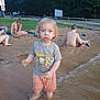 child, toddler, water, mud, beach, sand, boy, person, outdoor, nature, trees, water_play, summer, curly_hair, shirtless, shorts, parked_cars, road_sign, woman, blanket