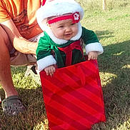 Zella is registered to the contest to win money with this photo: baby, child, person, adult, outdoor, grass, holiday_clothing, santa_hat, gift_bag, red, green, festive, smiling, happy, holding, sunlight, casual_clothing, wooden_structure, nature, daytime