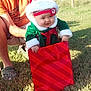 adult, baby, casual_clothing, child, daytime, festive, gift_bag, grass, green, happy, holding, holiday_clothing, nature, outdoor, person, red, santa_hat, smiling, sunlight, wooden_structure