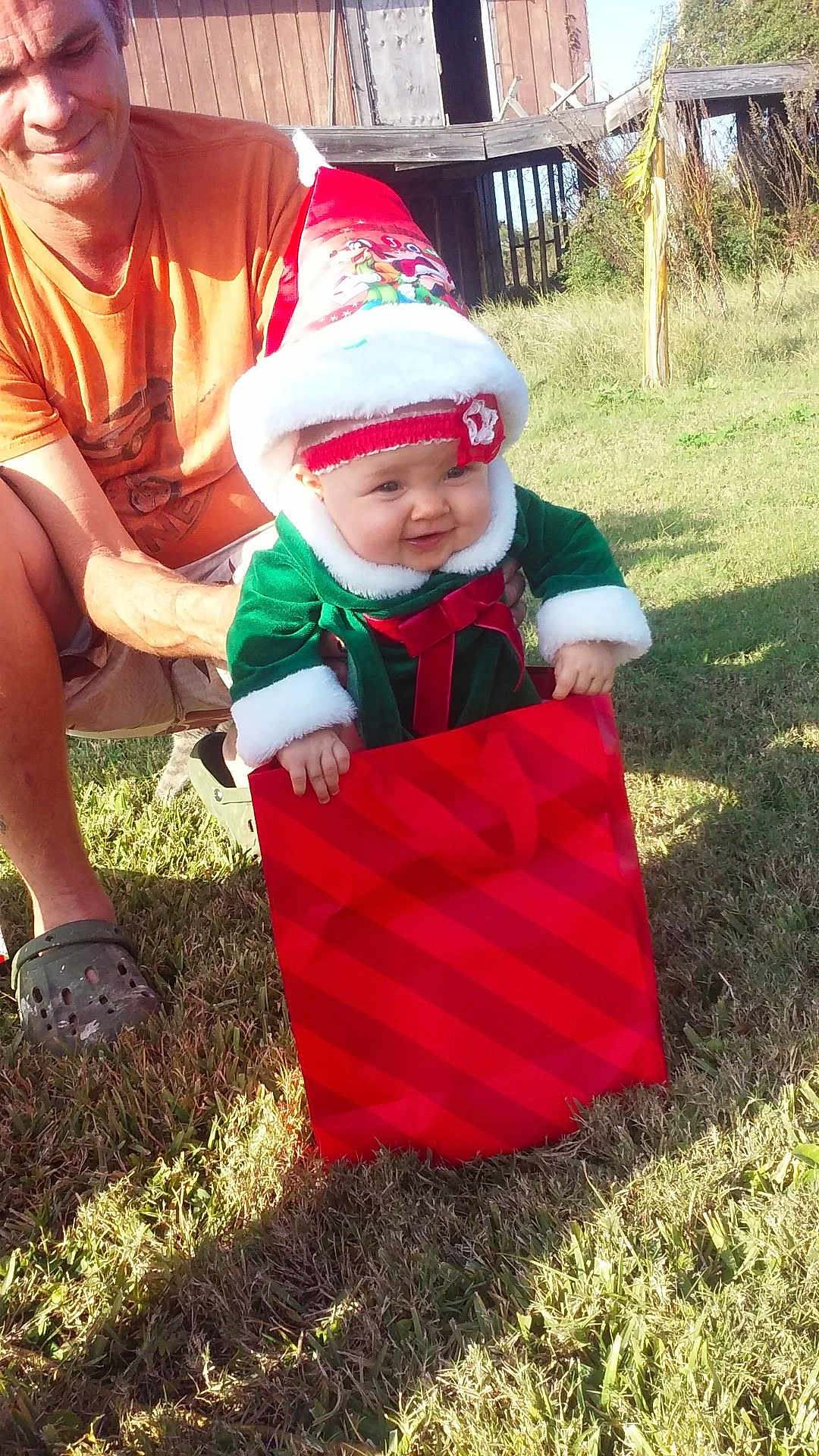 Zella is registered to the contest to win money with this photo: baby, child, person, adult, outdoor, grass, holiday_clothing, santa_hat, gift_bag, red, green, festive, smiling, happy, holding, sunlight, casual_clothing, wooden_structure, nature, daytime