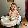 baby, child, high_chair, cupcake, candle, number_candle, whipped_cream, sprinkles, birthday, indoor, wood_floor, painting, wall, curtains, table, foot, face, person, celebration, food