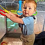 Luca joined the competition — help win amazing prizes! baby, child, playpen, toy, standing, blue_shirt, gray_shorts, blanket, pacifier, indoor, furniture, couch, wood_paneling, rings, face, person, home, curious, soft_lighting, floor
