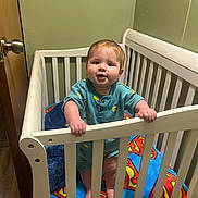 Luca joined the competition — help win amazing prizes! baby, crib, blanket, superman_logo, standing, indoor, child, railing, wooden_door, green_wall, cute, infant, hand, feet, clothing, smiling, face, room, furniture, portrait