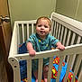 baby, crib, blanket, superman_logo, standing, indoor, child, railing, wooden_door, green_wall, cute, infant, hand, feet, clothing, smiling, face, room, furniture, portrait
