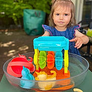 Mya a rejoint le concours — aidez-le/la à gagner de superbes lots ! toddler, child, toy, water, plastic_basin, outdoor, greenery, denim_dress, play, colorful, yellow, blue, orange, red, summer, daylight, curious, face, person, young