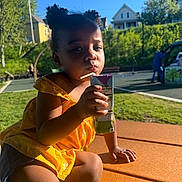 Aria joined the competition — help win amazing prizes! child, toddler, juice_box, yellow_dress, outdoor, sunlight, park, picnic_table, greenery, trees, houses, summer, cute, sitting, drinking, portrait, casual, daytime, person, playground