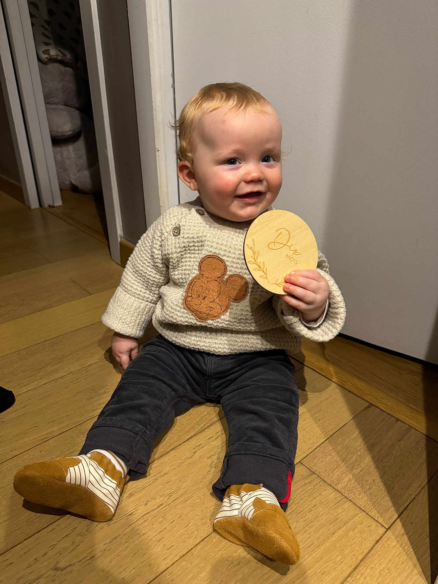 Lucas Bequin participe au concours pour gagner de l'argent avec cette photo : baby, child, sitting, floor, wooden_floor, sweater, mickey_mouse, plush_chair, doorway, pants, socks, shoes, holding, wooden_plaque, indoor, smiling, cute, happy, person, portrait