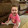 baby, child, smiling, sitting, pink_shirt, patterned_pants, carpet, couch, furniture, indoor, living_room, happy, person, face, footwear_free, cute, young_child, home, playful, infant