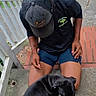 animal, black_dog, brick, candid, cap, casual_clothing, concrete, daytime, dog, grass, kneeling, outdoor, person, pet, playful, porch, railing, shorts, sidewalk, summer