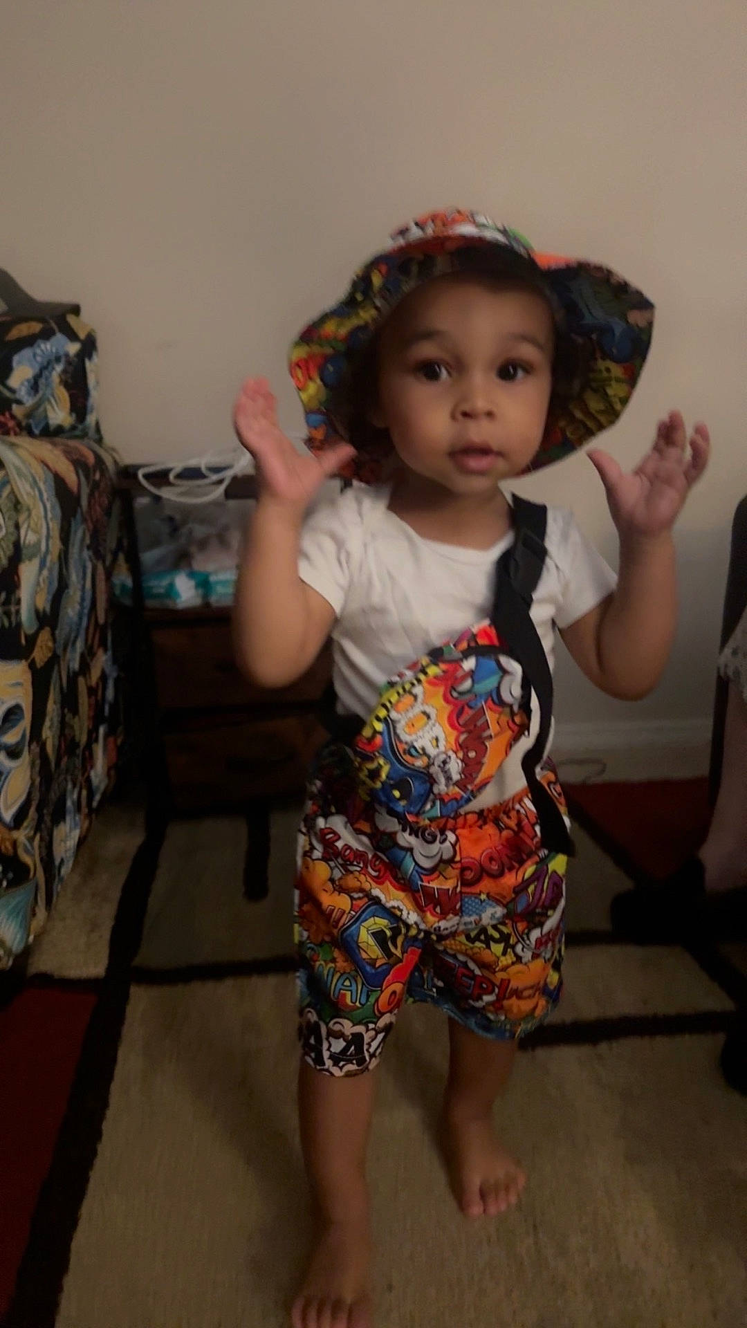 Amaro is registered to the contest to win money with this photo: barefoot, bucket_hat, carpet, child, colorful_clothing, curious, expression, furniture, hat, home, indoor, patterned_clothing, person, portrait, room, shorts, standing, toddler, white_tshirt, young_child