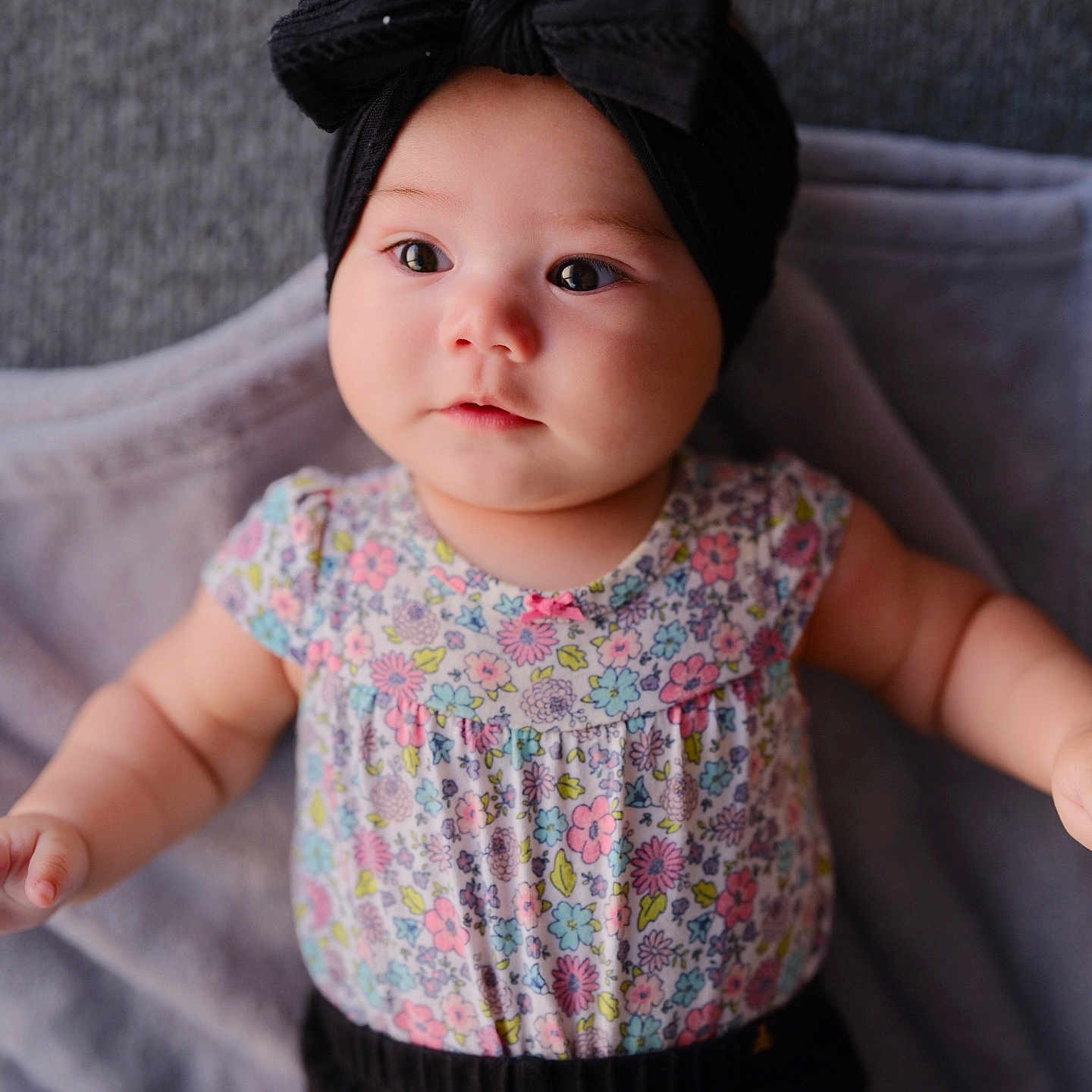 Ava Angeline is registered to the contest to win money with this photo: adorable, baby, black_pants, blanket, bow, child, chubby_cheeks, closeup, cute, expression, floral_clothing, headband, indoors, infant, lying_down, person, portrait, skin, soft_lighting, young_child