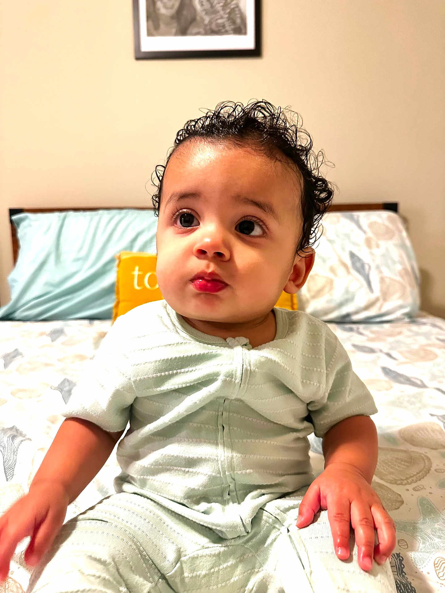 Dylan is registered to the contest to win money with this photo: baby, child, indoor, bed, pillow, curly_hair, light_green_clothing, hand, face, eyes, expression, portrait, infant, sitting, cozy, soft_lighting, bedroom, patterned_sheet, yellow_pillow, cute
