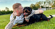 Zola participe au concours pour gagner de l'argent avec cette photo : affection, casual_clothing, clouds, dog, fence, grass, greenery, happy, kiss, man, nature, outdoor, pet, playful, relaxing, rottweiler, sky, summer, tattoos, tongue_out