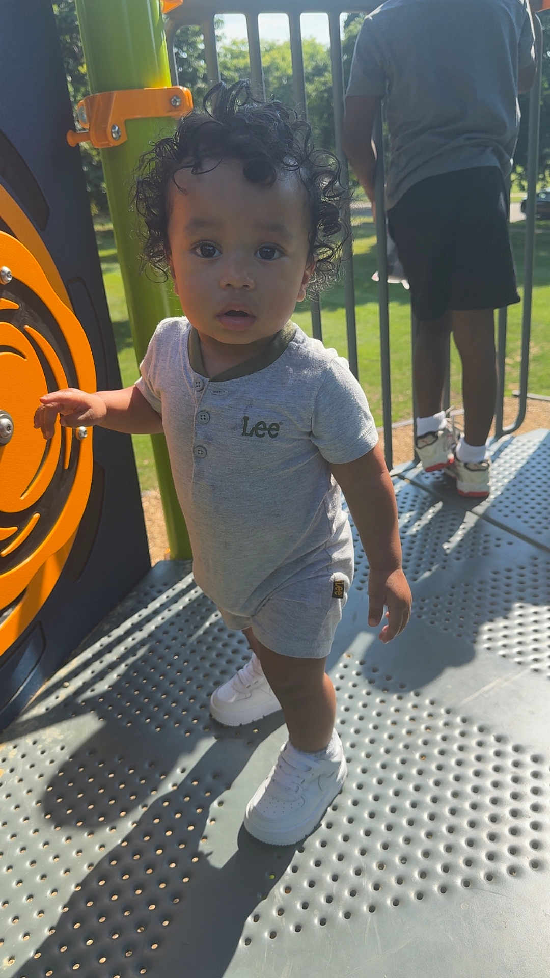 Jovani joined the competition — help win amazing prizes! toddler, child, curly_hair, playground, outdoor, sunlight, grey_outfit, white_sneakers, person, metal_floor, play_equipment, shadow, greenery, summer, casual_clothing, standing, daytime, background_person, park, curious_expression