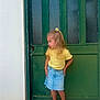 Abby participe au concours pour gagner de l'argent avec cette photo : child, girl, yellow_top, denim_skirt, flower_hair_clip, blonde_hair, green_door, standing, sandals, curious, outdoor, side_view, wall, footwear, portrait, casual_clothing, hands_on_hips, young_child, daylight, tile_floor