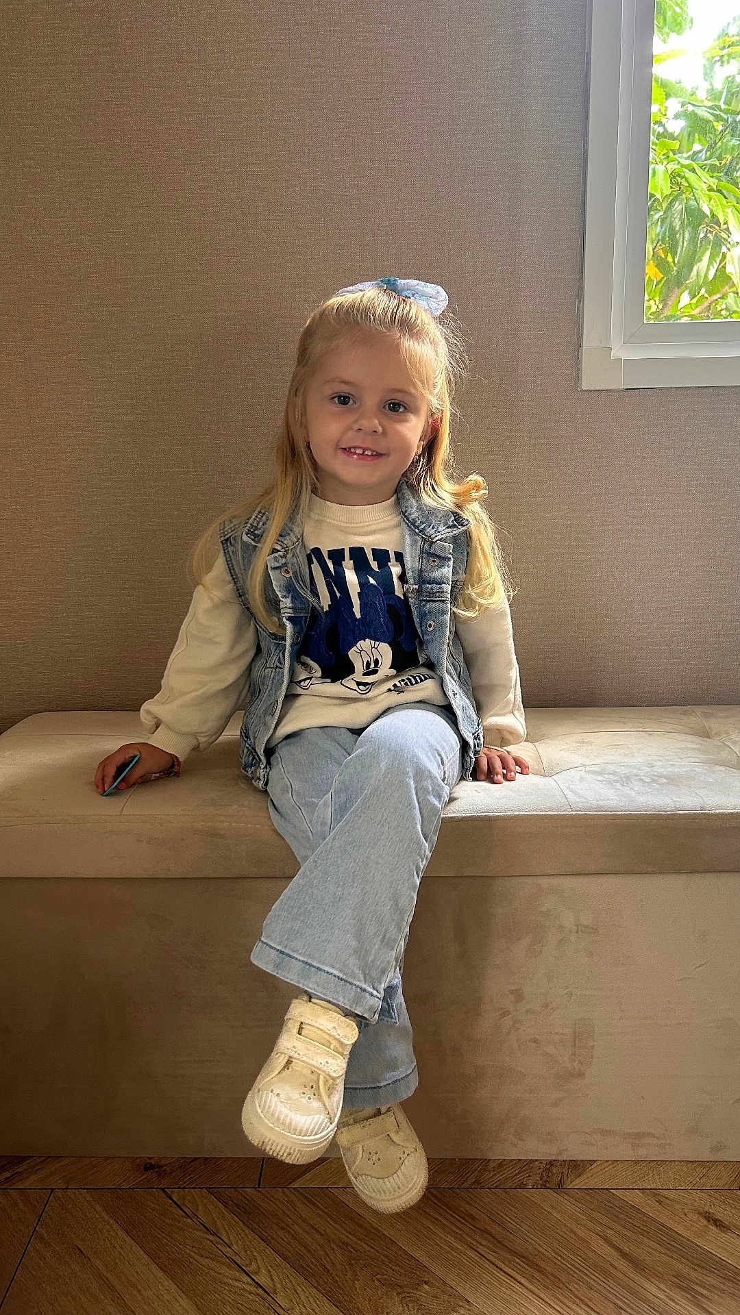 Abby participe au concours pour gagner de l'argent avec cette photo : child, girl, smiling, indoor, window, denim, sweater, blonde, hair_bow, sneakers, bench, wooden_floor, casual_clothing, portrait, happy, seated, light, natural_light, cute, home