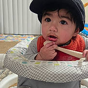 Ali joined the competition — help win amazing prizes! baby, child, infant, cap, bib, spoon, baby_seat, clothing, face, person, indoor, carpet, curtain, toy, expression, cute, young_child, sitting, home, curious