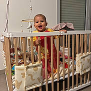 Charlotte participe au concours pour gagner de l'argent avec cette photo : baby, child, crib, mobile, toy, stuffed_animal, pajamas, smile, happy, indoor, wooden_bars, playpen, bedding, blanket, window_blind, floor_tile, person, hand, dimple, nursery