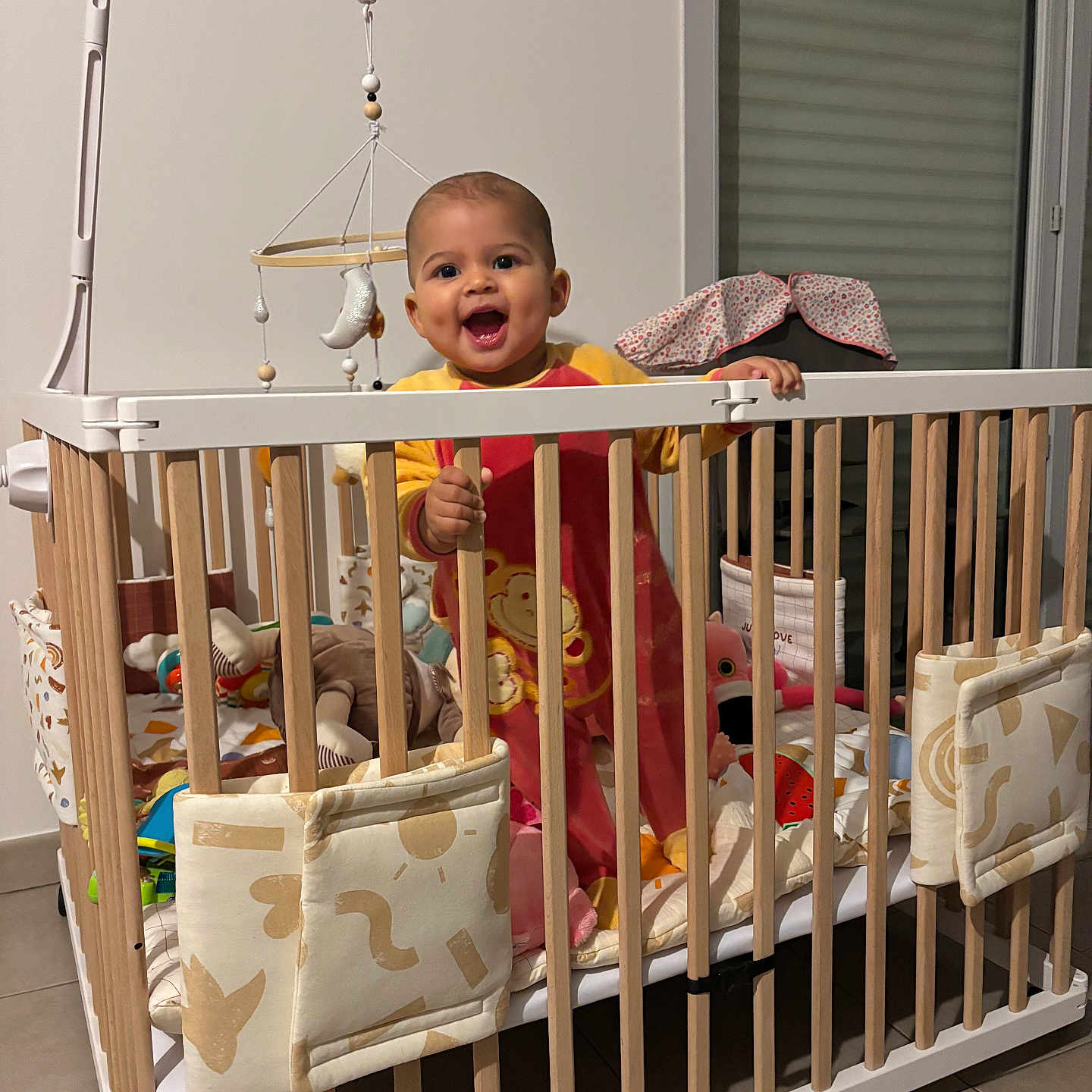 Charlotte participe au concours pour gagner de l'argent avec cette photo : baby, bedding, blanket, child, crib, dimple, floor_tile, hand, happy, indoor, mobile, nursery, pajamas, person, playpen, smile, stuffed_animal, toy, window_blind, wooden_bars