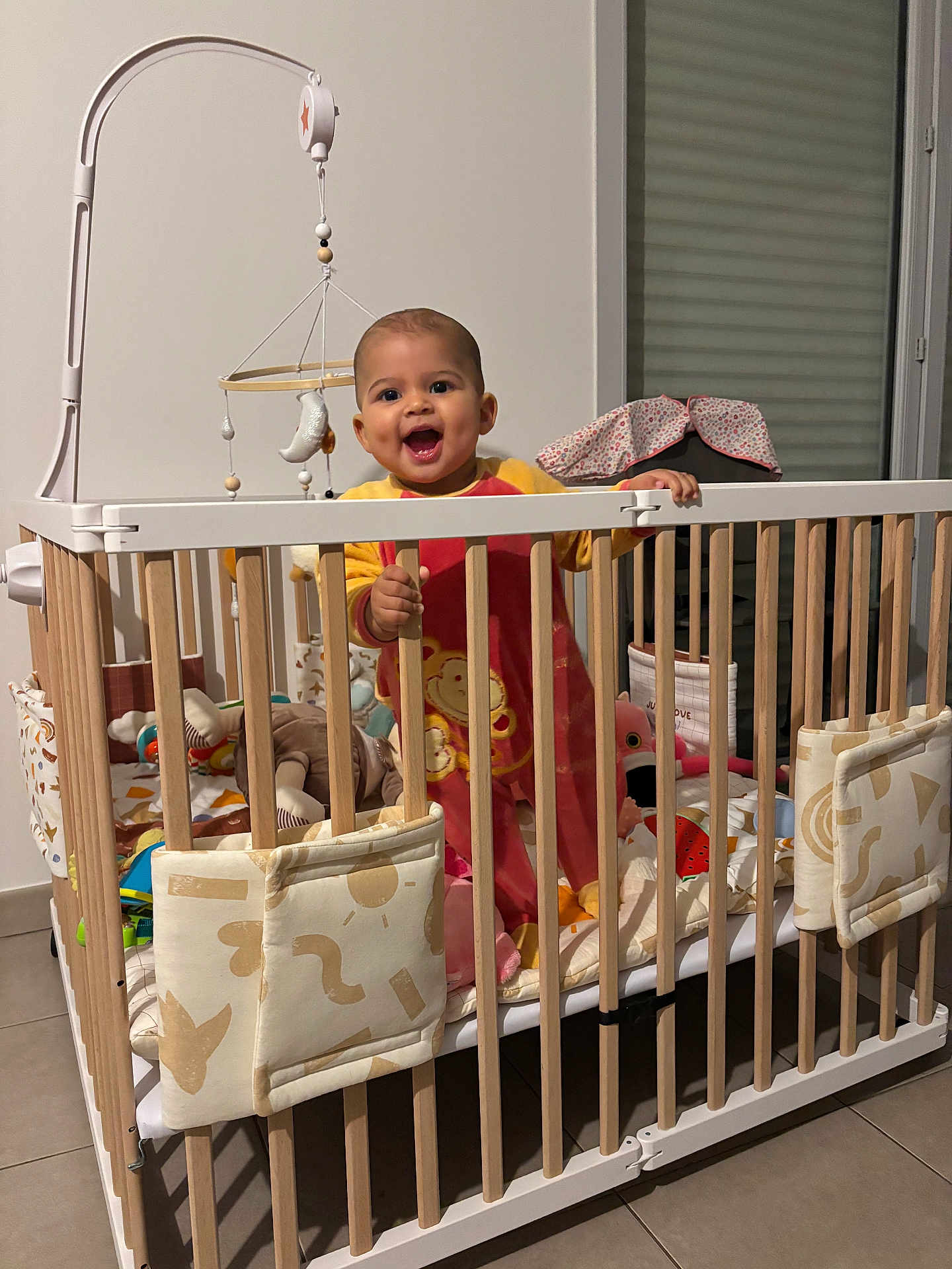 Charlotte participe au concours pour gagner de l'argent avec cette photo : baby, child, crib, mobile, toy, stuffed_animal, pajamas, smile, happy, indoor, wooden_bars, playpen, bedding, blanket, window_blind, floor_tile, person, hand, dimple, nursery