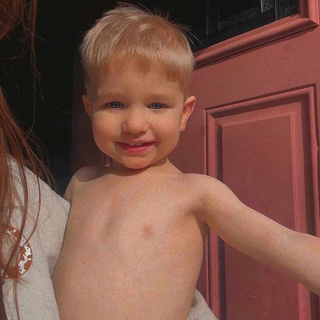 Remmington joined the competition — help win amazing prizes! adult_hand, arm, blond_hair, child, closeup, cute, door, face, happy, holding, indoors, person, portrait, red_door, shirtless, skin, smiling, sunlight, toddler, young_child