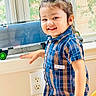 Arshan is registered to the contest to win money with this photo: child, toddler, smiling, portrait, indoors, window, sunlight, toy_vehicle, plaid_shirt, blue_clothing, short_sleeves, ponytail, hair_bun, standing, hand, face, teeth, playroom, electrical_outlet, home