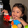Arshan is registered to the contest to win money with this photo: child, toddler, smiling, portrait, toy_train, toy_vehicles, playtime, indoors, table, water_bottle, dark_background, black_chair, colorful_toys, red_toy, yellow_toy, green_toy, blue_toy, long_hair, happy, close_up