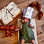 baby, infant, chef_hat, paper_hat, flour, whisk, eggs, baguette, croissants, wood_floor, towel, green_romper, food, baking, cute, lying_down, face, hands, legs, floor