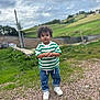blue_jeans, casual_clothing, child, cloudy_sky, curly_hair, daylight, field, grass, gravel, green_shirt, nature, outdoor, person, portrait, rural, sky, standing, striped_shirt, toddler, white_sneakers