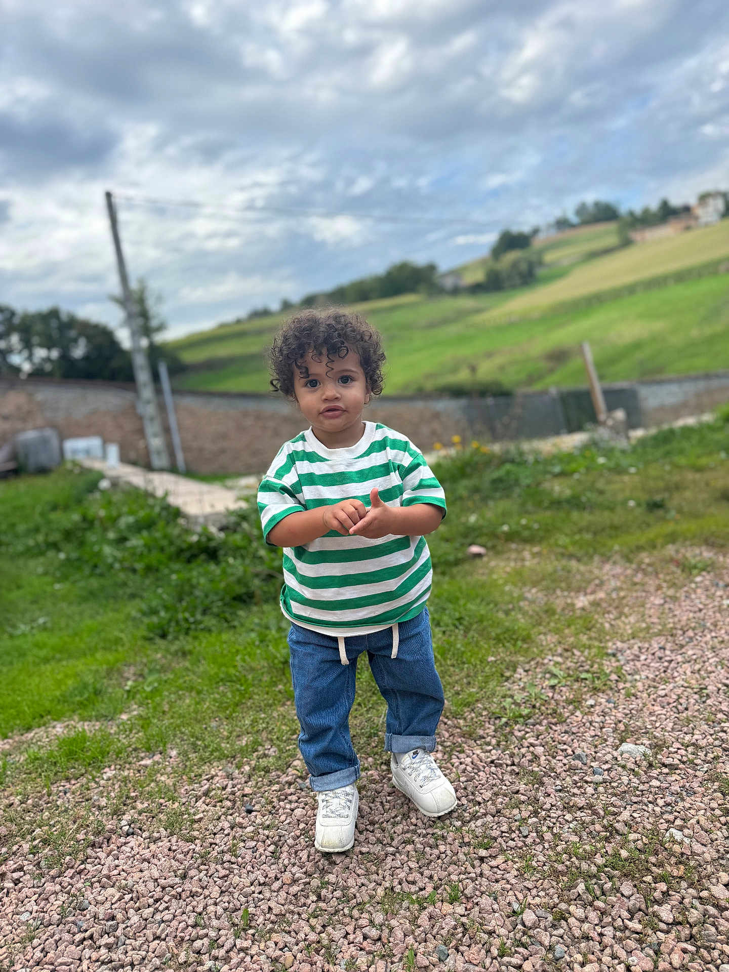 Khelis participe au concours pour gagner de l'argent avec cette photo : child, toddler, curly_hair, green_shirt, striped_shirt, blue_jeans, white_sneakers, gravel, grass, outdoor, sky, cloudy_sky, field, nature, person, standing, casual_clothing, daylight, portrait, rural
