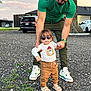 toddler, child, adult, walking, sunglasses, bear_shirt, green_shirt, green_pants, sandals, sneakers, gravel, outdoor, tree, car, supporting, holding_hands, casual_clothing, daylight, person, portrait