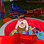 child, toddler, ball_pit, slide, outdoor, playground, night, string_lights, grass, toy, colorful, red_slide, blue_toy, brown_pants, bear_shirt, playing, multicolored_balls, person, fun, recreation