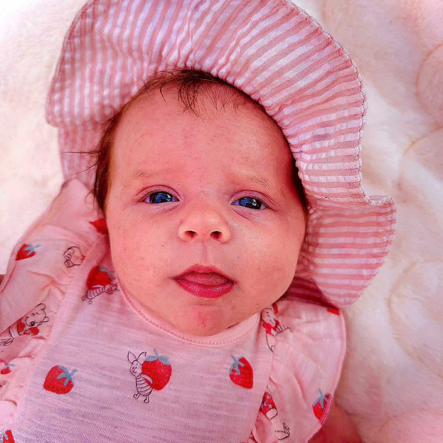 Amelia joined the competition — help win amazing prizes! apparel, baby, bear_pattern, blue_eyes, child, closeup, cute, face, fluffy_blanket, headwear, infant, lying_down, newborn, pink_clothing, pink_hat, portrait, soft_texture, strawberry_pattern, striped_hat, tongue_out