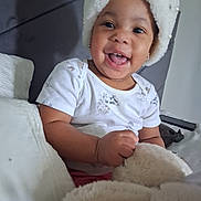 Arielle is registered to the contest to win money with this photo: baby, child, toddler, smiling, happy, portrait, indoors, sitting, winter_hat, fluffy_hat, teddy_bear, stuffed_animal, white_clothing, short_sleeves, earrings, necklace, couch, blanket, bedroom, close_up